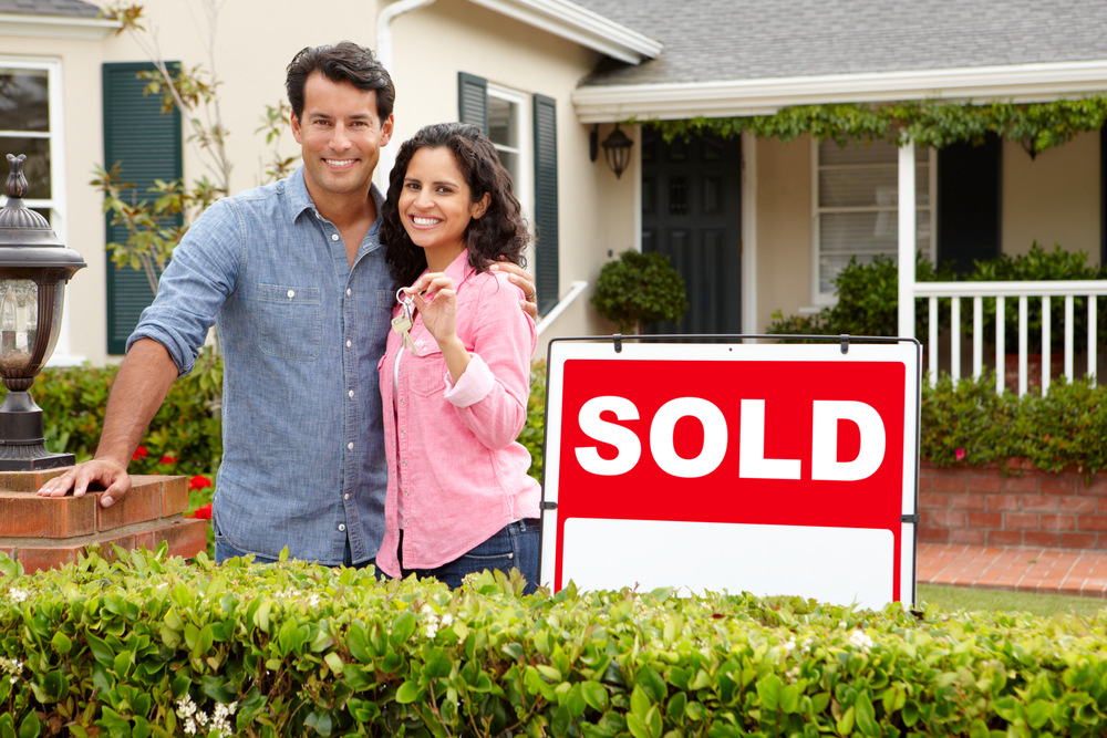 couple hugging smiling holding keys in front of a house with sold sign